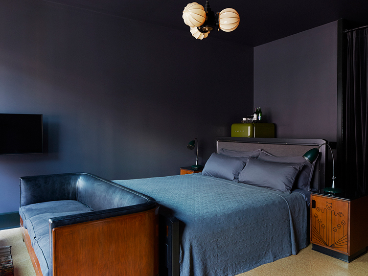 bedroom with midnight blue walls, blue bed and blue leather couch with deco wood side tables.